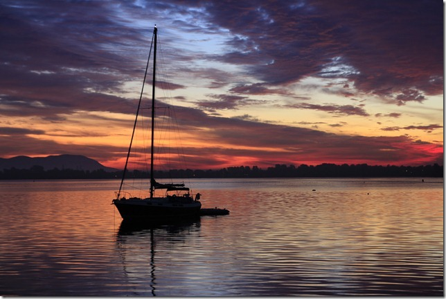 Silhouette d’Évasion au mouillage sur une eau calme, sous un ciel de coucher de soleil pourpre et orangé