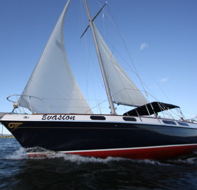 Évasion sous voile, coque bleu marine et ligne de flottaison rouge, génois et grand-voile gonflés sous un ciel bleu